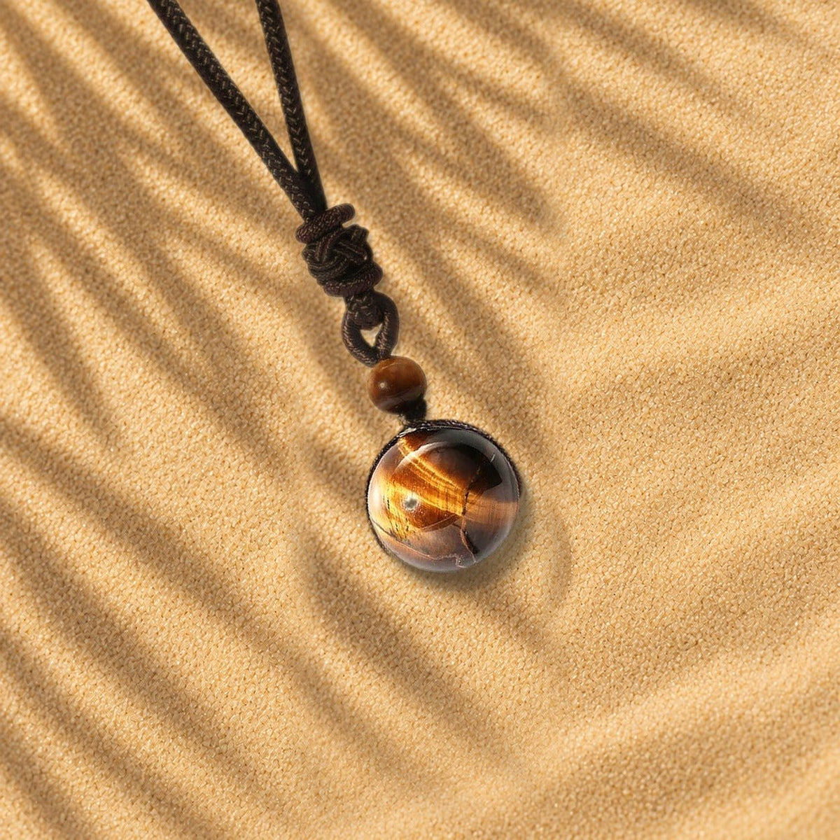 Collier œil de tigre Orbe, pendentif sphère en pierre naturelle posé sur le sable